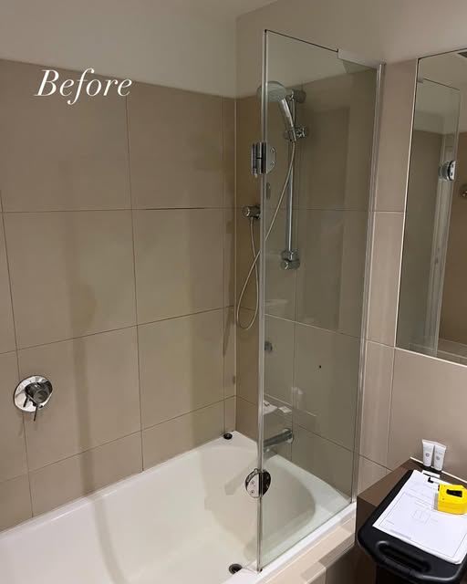 A dated framed shower screen over a bath in a beige tiled bathroom, before the work